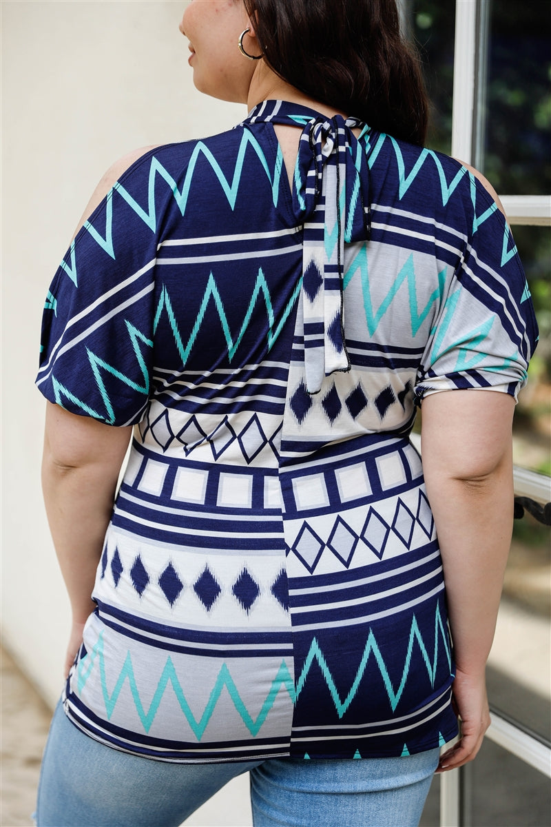 A stylish plus-size woman wearing a navy and mint polka dot cold shoulder top with short sleeves and a back self-tie detail.