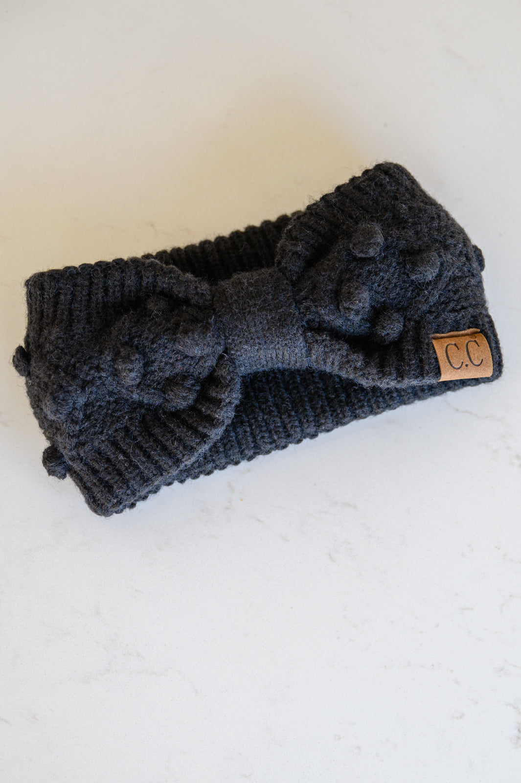 Black Pom Knit Headwrap featuring a bow front and pom pom detailing, made from soft ribbed knit material.