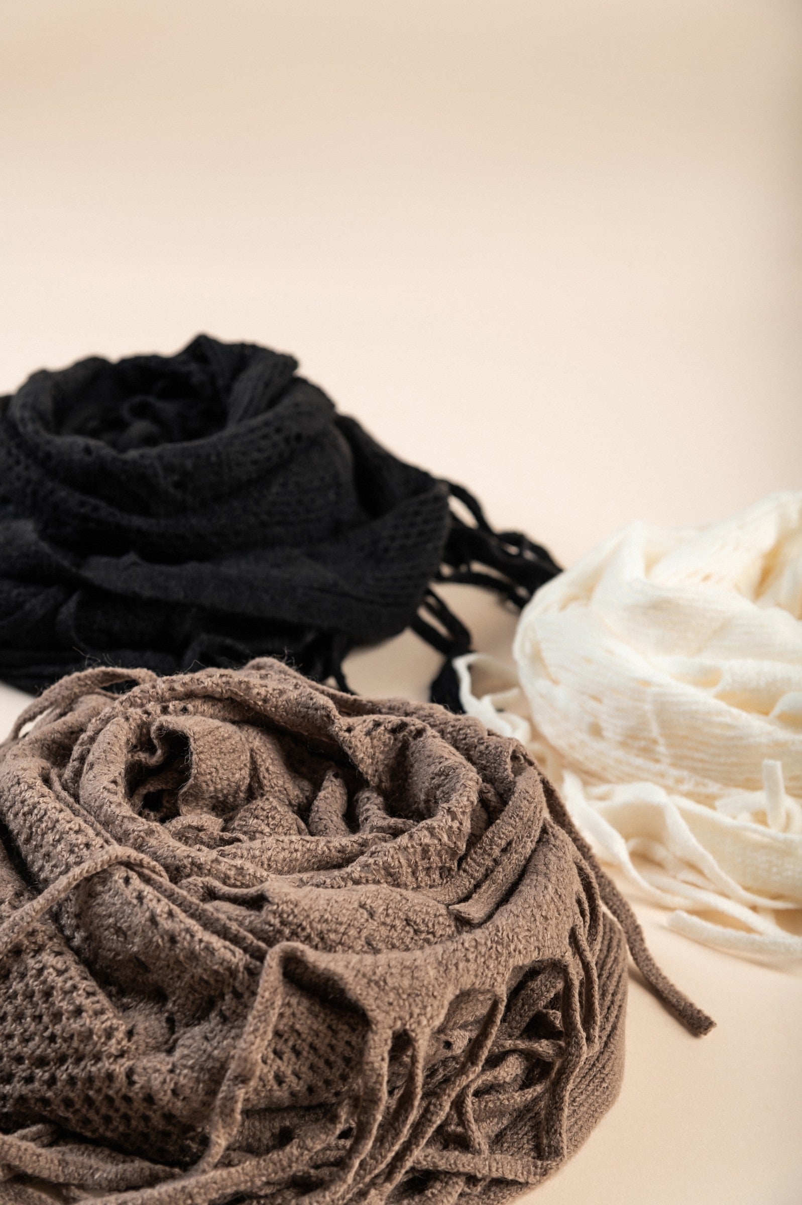 Set of 3 knitted scarves in white, black, and beige with fringes, showcasing their triangular shape and soft texture.