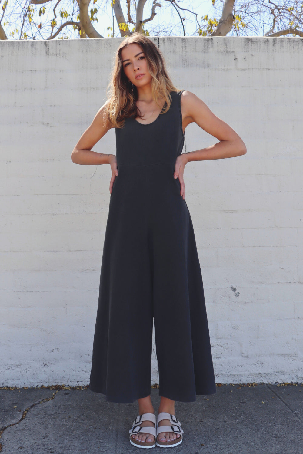 A stylish Sunset Jumpsuit featuring a waist-defining seam, wide legs, and v-neck detailing, available in Washed Black, Sand, and Persimmon colors.