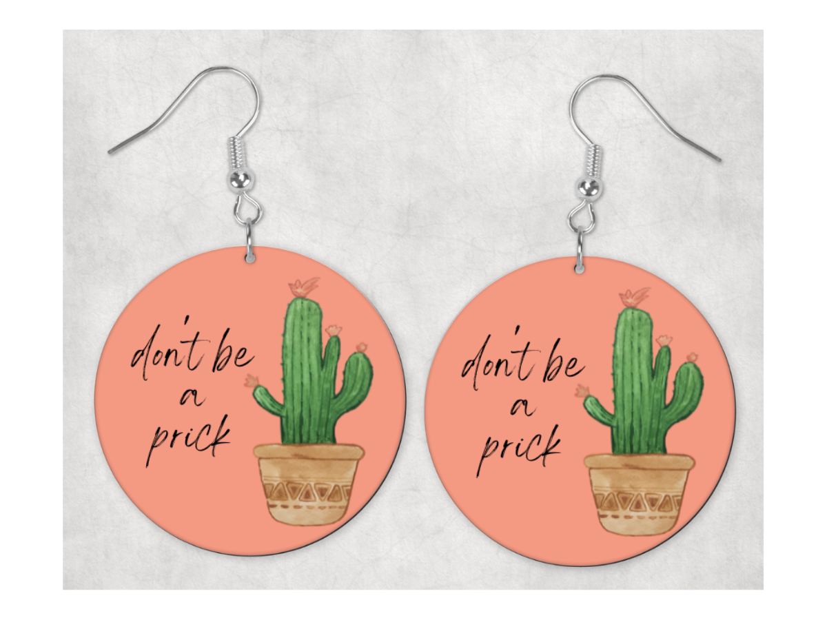 A pair of 1-inch circle earrings with a humorous phrase 'Don't Be A Prick' on a hook ear wire, showcasing a playful and stylish design.