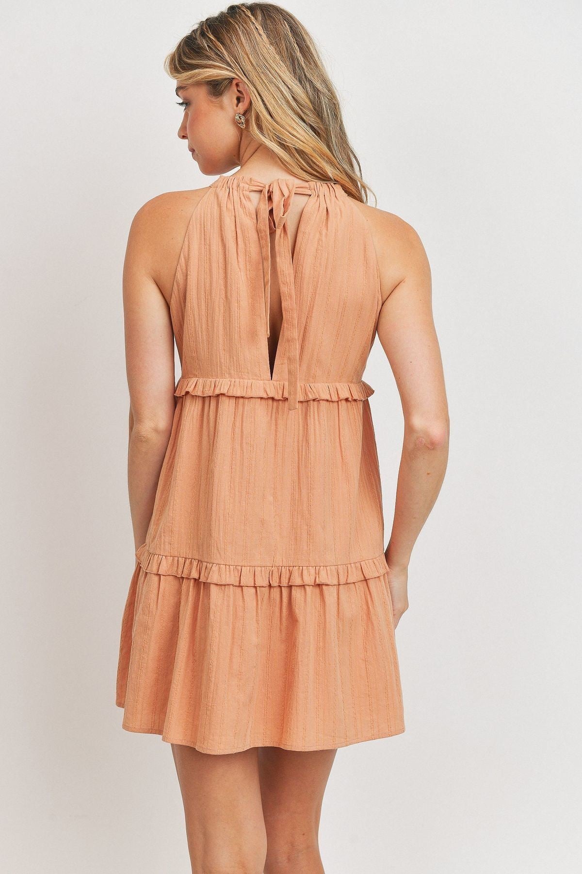 A stylish blush tiered dress made from textured woven fabric, featuring a halter neck and ruffled layers, perfect for summer occasions.