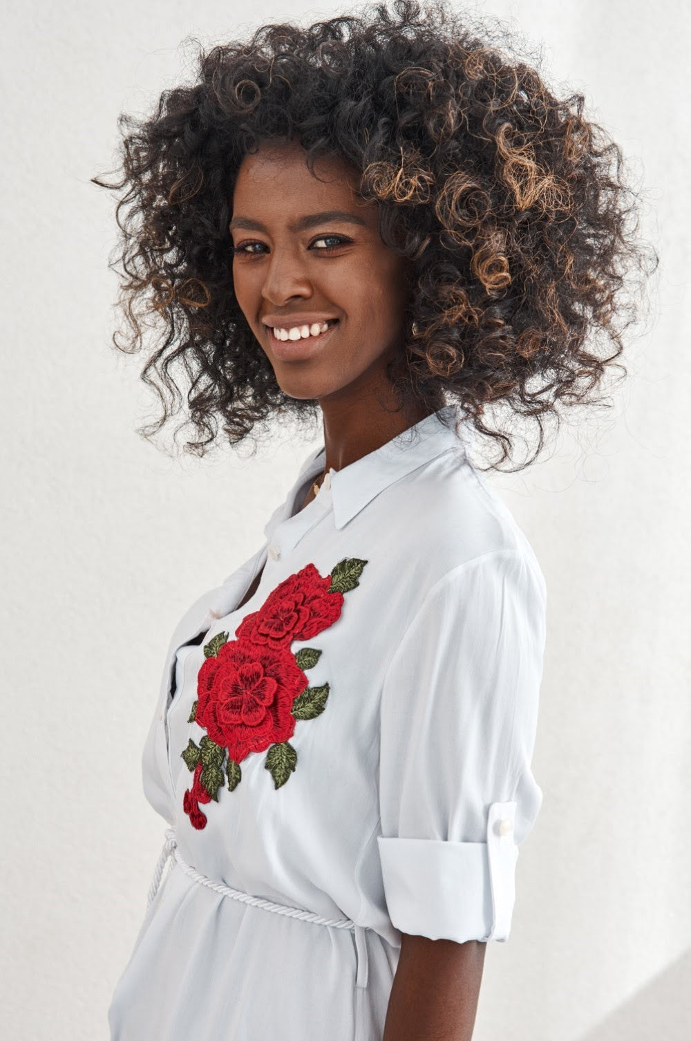 A stylish white shirt dress featuring beautiful embroidered flowers, buttoned from collar to waist with rolled-up sleeves and a tied belt.